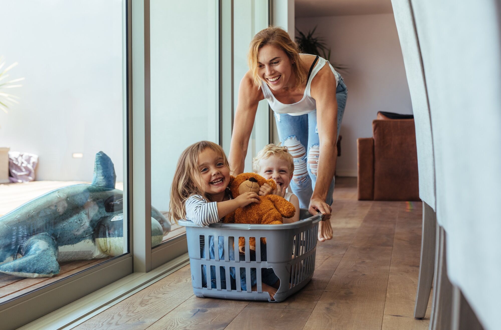 A women pushing two laughing children in a laundry basket on the ground.