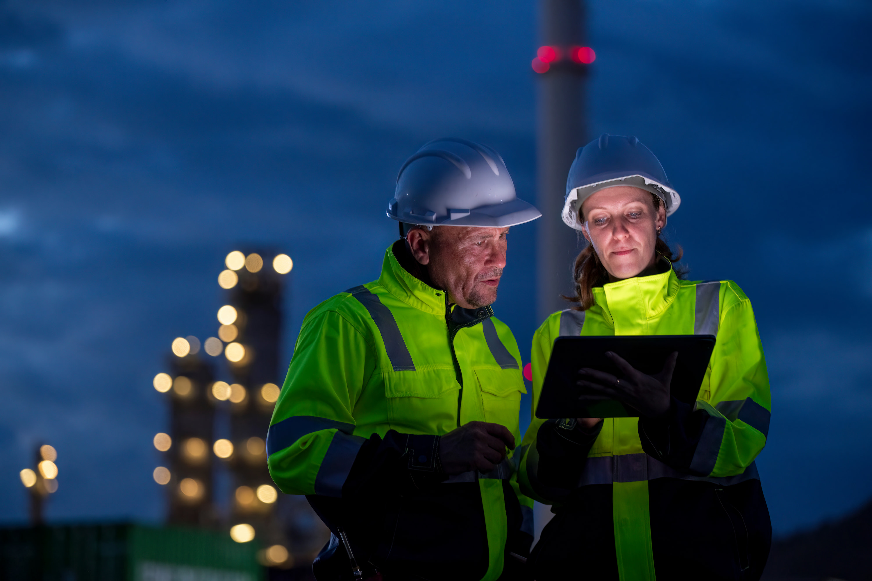 Two workers wearing hardhats and safety vests using a tablet at night.